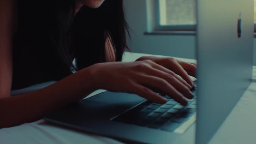 Woman smiles while lying on her bed and typing on a computer