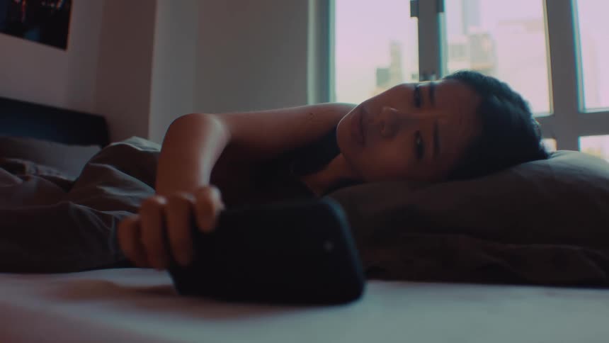 Person lying on a bed reaches toward a smartphone beside the pillow