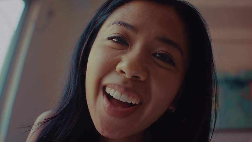 A young woman sits indoors at midday smiling brightly as she waves excitedly during a video conference call sunlight fills the room giving the scene a warm lively feel while she greets the caller with enthusiasm