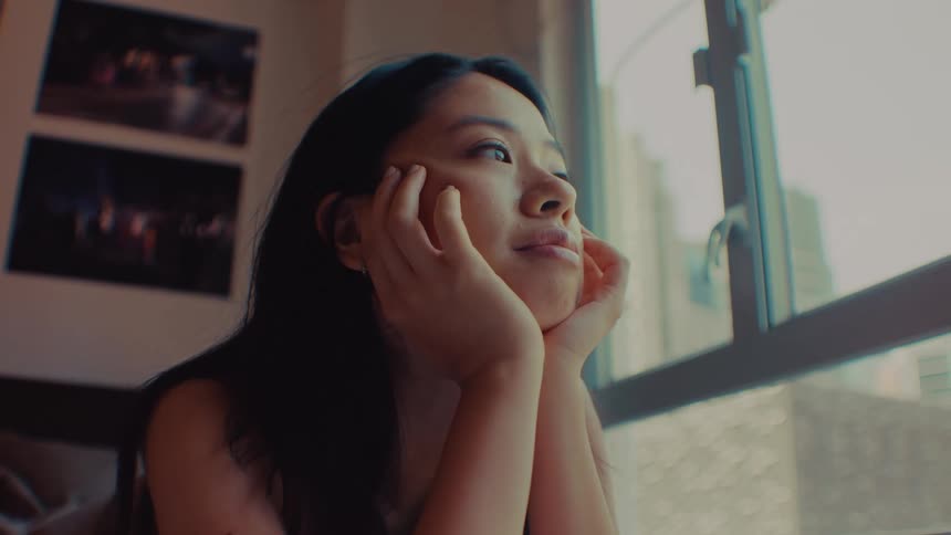 A woman rests her chin on her hands and gazes out a window in a dimly lit room looking forward with a thoughtful mood