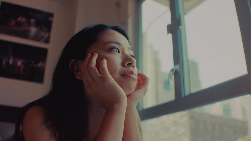 A woman rests her chin on her hands and gazes out a window in a dimly lit room looking forward with a thoughtful mood
