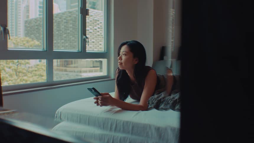 An asian woman lies on a bed and looks at a smartphone by a bright window in a dimly lit room looking forward with a calm mood