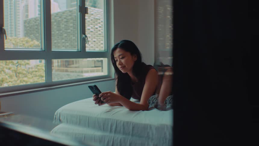 An asian woman lies on a bed and looks at a smartphone by a bright window in a dimly lit room looking forward with a calm mood