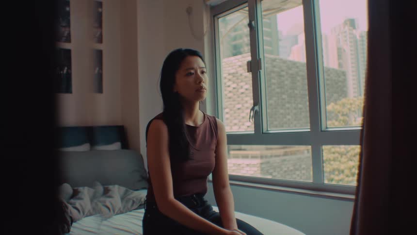 A woman in a sleeveless top sits on a white bed and gazes toward a window in a dimly lit room looking forward with a sad pensive mood