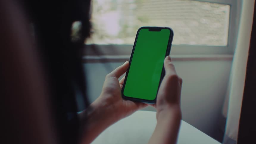 A woman sits at a white table and holds a smartphone with a green screen and she uses her thumb to tap the center of the display while looking at the device near a window with sunlight coming through the blinds