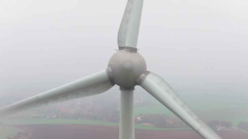 The video shows a closeup aerial shot of a large wind turbine with three blades rotating slowly against a foggy overcast sky as the camera pulls back it reveals a rural landscape below with patches of brown fields and a small hazy village in the dist