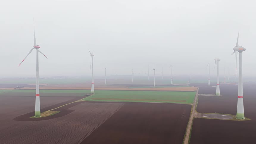An aerial drone shot captures several tall wind turbines standing in vast flat agricultural fields under a thick gray mist