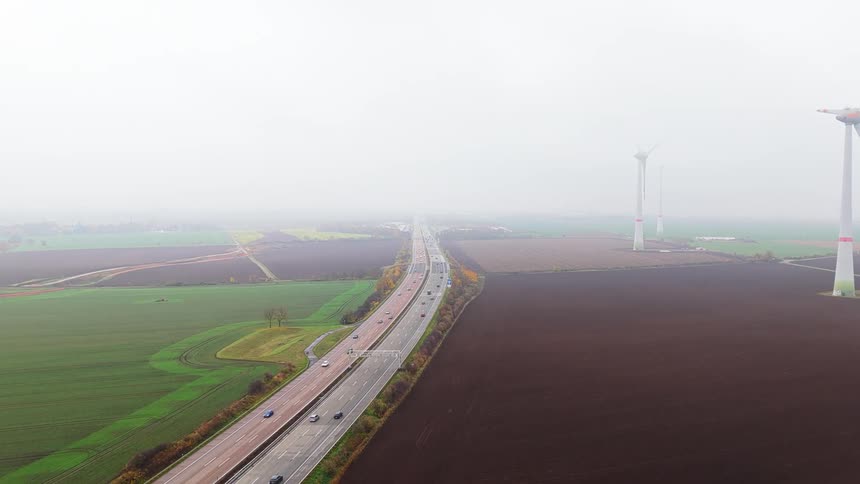 A busy highway in between a green grass field and a windmill farm under a foggy sky