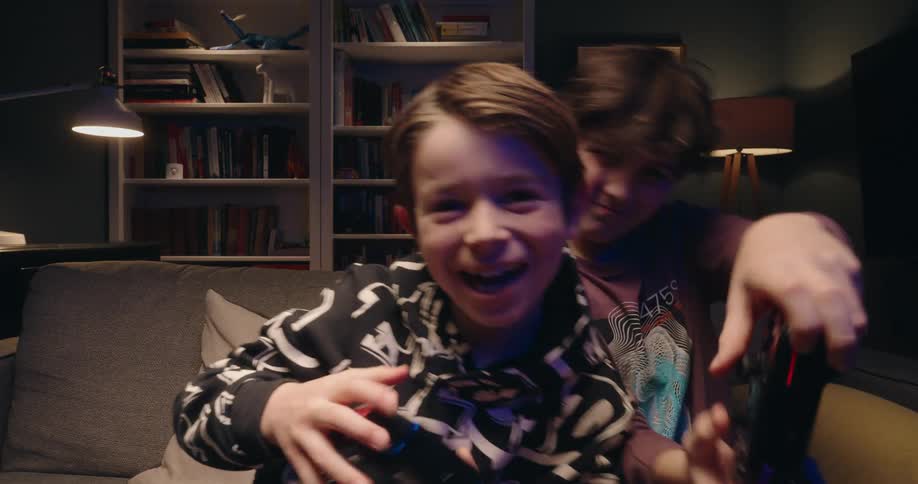Two young boys are sitting on a gray couch in a dimly lit room energetically playing a video game while looking directly at the camera they are laughing and moving their controllers with excitement framed by a bookshelf and a warm lamp in the backgroun