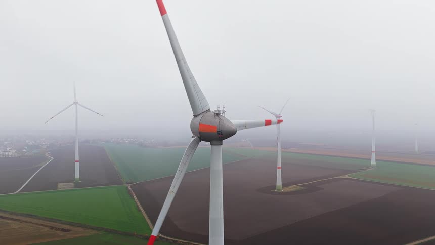 An aerial shot captures a large wind turbine spinning its redtipped blades above a vast patchwork of farmland under a grey foggy sky