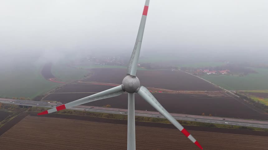 A close up of a windmill with a busy highway and a soil and grass field in the background under a foggy sky