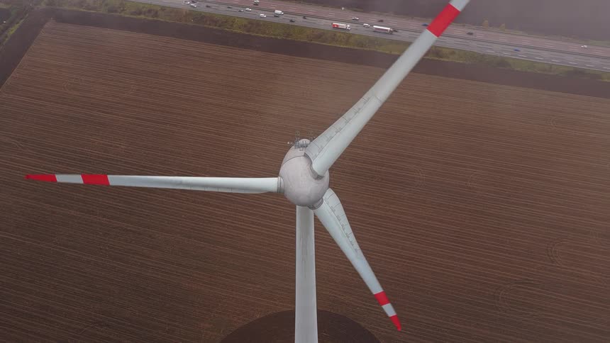 An aerial high angle shot shows a large white wind turbine with redtipped blades spinning slowly over a brown field next to a busy highway