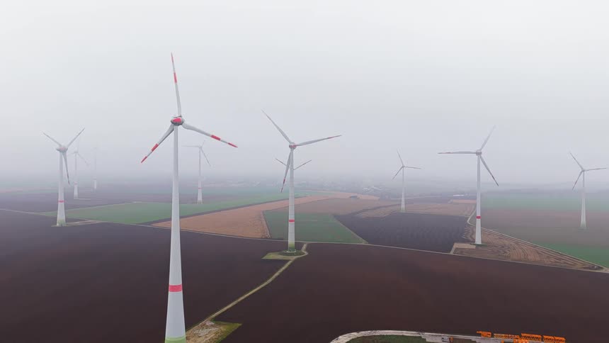 An aerial drone shot pans over a vast wind farm with several tall turbines on a misty day