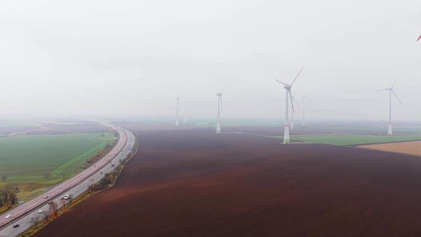 Wind turbines spread across agricultural fields with a highway alongside under an overcast sky