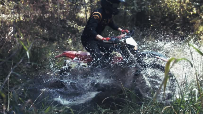 A rider on a red dirt bike powers through a flooded forest trail sending a massive spray of water and mud flying into the air as they navigate the rough terrain