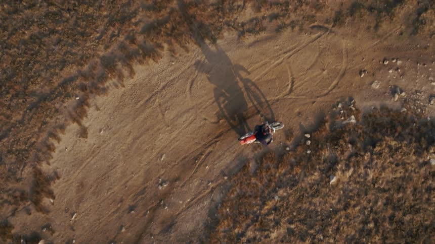 From a high topdown perspective a motocross rider sits stationary on a dirt trail casting a long sharp shadow in the late afternoon sun