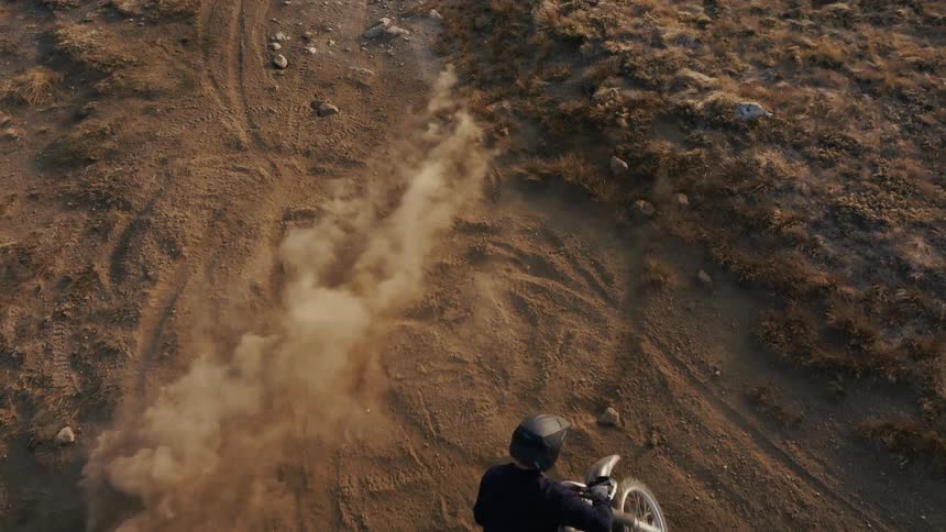 A motorcyclist wearing a black helmet and gear rides a red dirt bike across a dusty arid landscape kicking up a large plume of brown earth as seen from an overhead aerial perspective