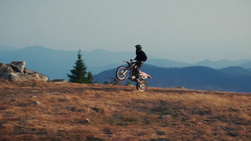 The video features a motorcyclist performing maneuvers on a dirt bike across a highaltitude mountainous ridge in the first segment the rider pops a wheelie while traveling along the crest of the hill against a backdrop of distant hazy blue mountain r