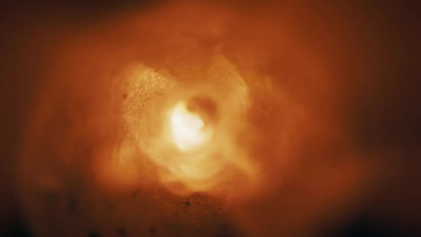 A glowing orange light radiates from the center of a dark textured tunnellike structure wisps of smoke or vapor drift through the narrow passage as the light begins to fade into shadow