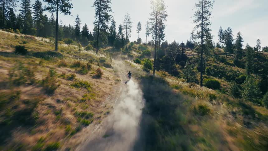 The video shows a highspeed fpv drone chase of a motorcyclist racing down a dusty trail in a hilly forested landscape a thick plume of white dust trails behind the rider catching the bright sunlight as the camera dives through the scattered pine trees