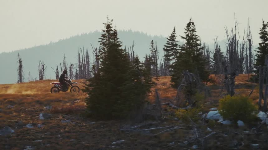 A man wearing black gear rides a dirt bike across a dry grassy mountain ridge during sunset and the camera pans to follow the rider
