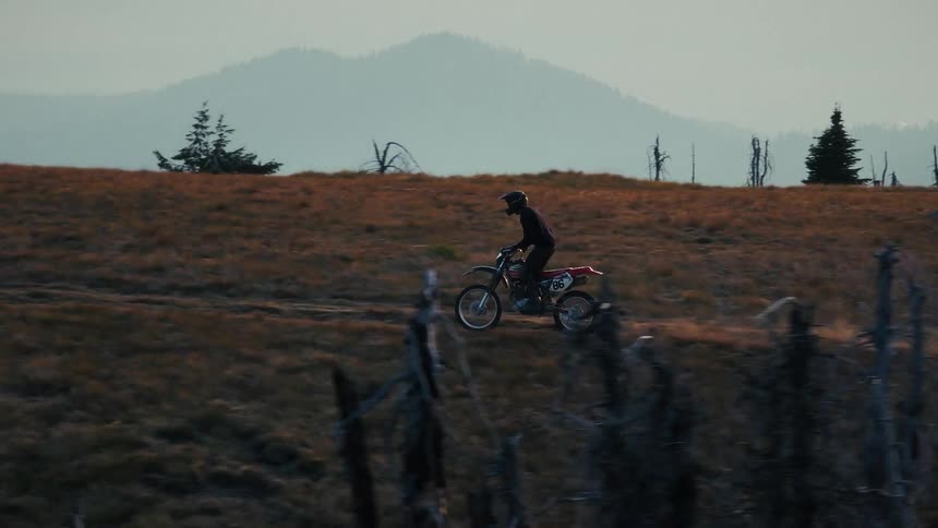 A lone motorcyclist clad in black gear rides across a vast goldenhued mountain ridge during the muted light of dusk the rider traverses a rugged trail against a backdrop of distant peaks and scattered skeletal remains of burnt trees