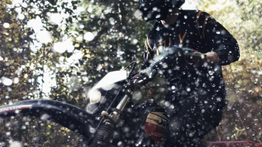 A man wearing a black helmet and jersey rides a dirt bike through a shallow body of water in a forest creating a large splash that fills the foreground as he moves toward the camera under bright sunlight filtering through the trees