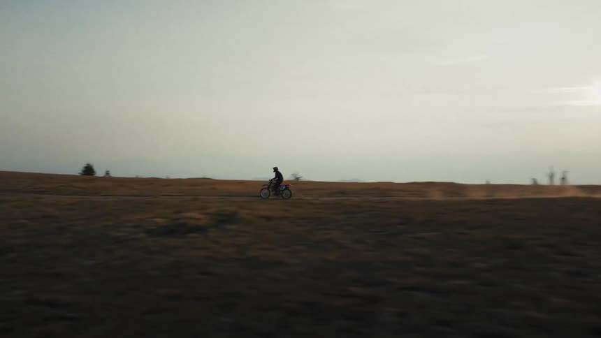 A lone fire lookout tower rises on a quiet grassy ridge at dusk as a single cyclist rides along a winding trail beneath it with hazy mountain layers stretching into the distance
