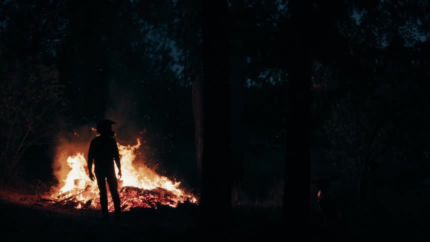 Person stands silhouetted against flames from a large wildfire in a dark forest at night