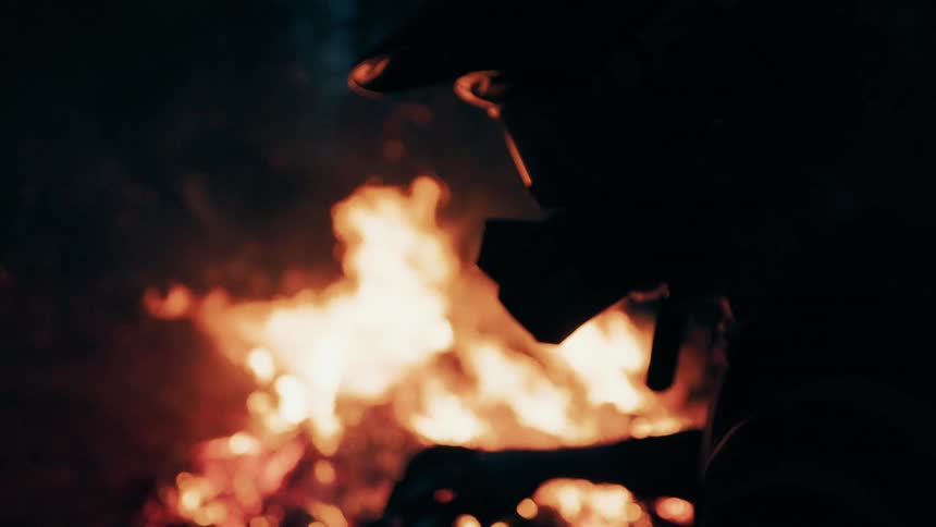 Silhouette of a person wearing a mask in front of a lit bonfire at night