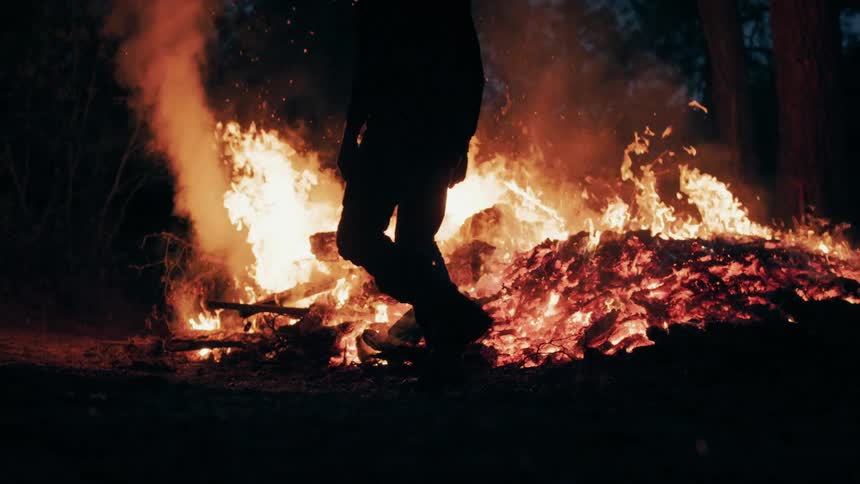 A silhouette of a person in dark clothing and boots walks past a large intense bonfire at night