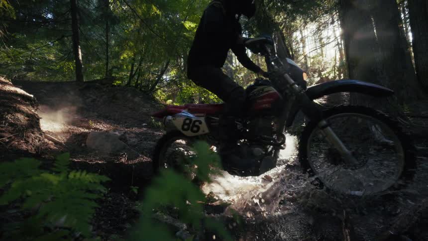 Motocross rider crossing a stream on a forest trail with water splashes lit by natural light