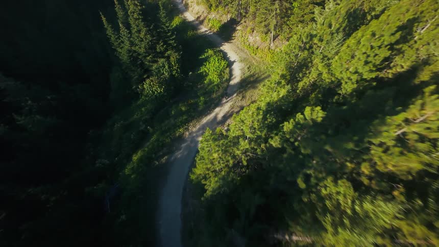 A drone follows a man riding a dirt bike down a winding mountain trail surrounded by dense evergreen forests under bright daylight capturing the descent from an overhead view into a close chase position