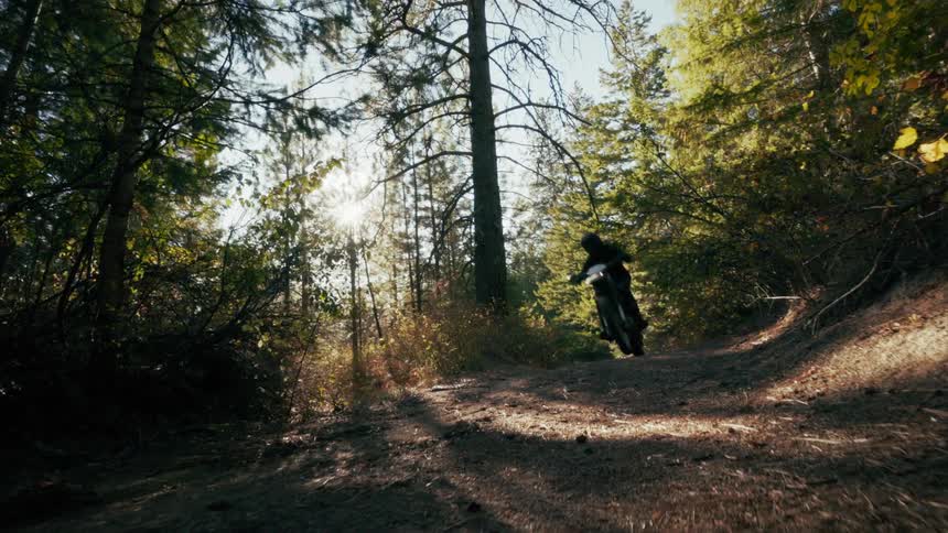 A dirt bike rider quickly travels across the screen from right to left kicking up a cloud of dust as they speed along a narrow forest trailthe sun shines brightly through the dense canopy of tall evergreen trees creating a strong lens flare and illum