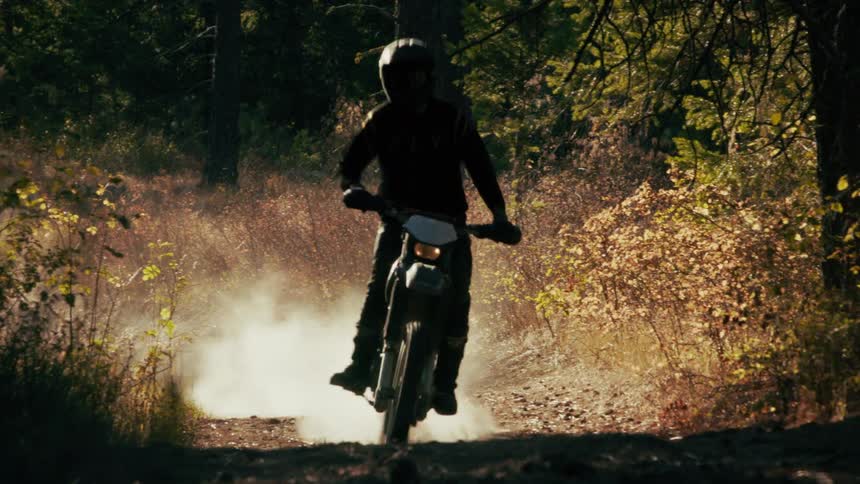 A dirt bike rider wearing a helmet and gear maneuvers down a sundrenched dusty trail through a wooded areathe bike kicks up a plume of dust as it approaches the camera eventually passing so close that the screen fades to black