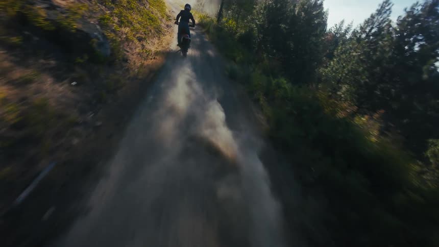 A motorcyclist rides down a dusty trail through a sun drenched mountain forest
