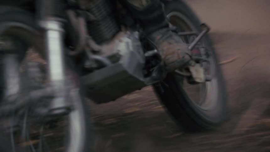 A motorcyclist wearing a black helmet and jersey weaves through a dense dark forest on a dirt path the camera captures a tight shaky shot as the bike kicks up a cloud of dust that quickly obscures the frame