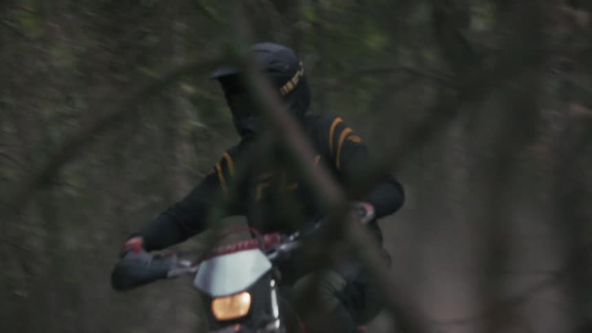 A man wearing a black helmet and a long sleeve jersey rides a motorcycle quickly through a dark forest on a dirt path as the camera follows his movement through the trees