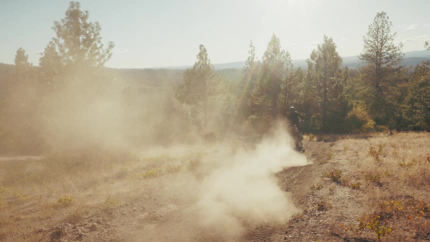 Motocross rider moving away along a dirt trail with dust rising in a wooded open area