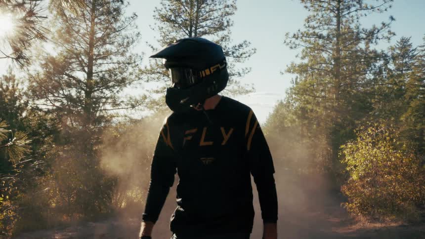 A person wearing a black and yellow motocross jersey and a matching helmet with goggles stands on a dirt path in a sunlit forest as dust swirls around them