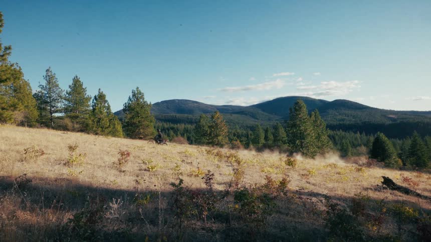 A motorcyclist rides a dirt bike across a dry grassy hillside kicking up a trail of dust against a backdrop of evergreen forests and rolling blue mountains under a clear afternoon sky