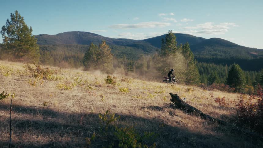 A motorcyclist rides across a grassy hillside kicking up dust with forested mountains in the background