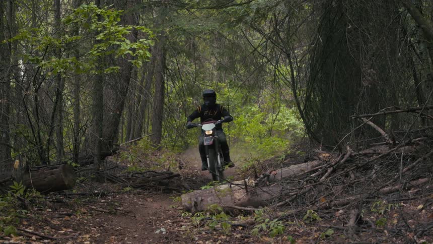 A dirt bike rider wearing black and yellow gear emerges from the shadows of a dense overgrown forestthe rider navigates a narrow dirt trail and kicks up a cloud of dust while passing quickly through the frame