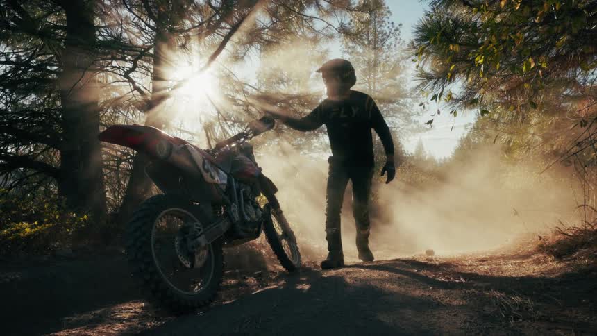 A person wearing a helmet and riding gear walks toward a dirt bike parked on a dusty trail in a sunlit forest