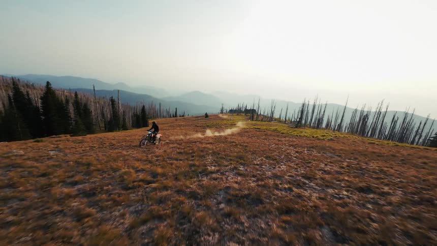 A person rides a dirt bike quickly across a high mountain ridge kicking up dust on a narrow trail