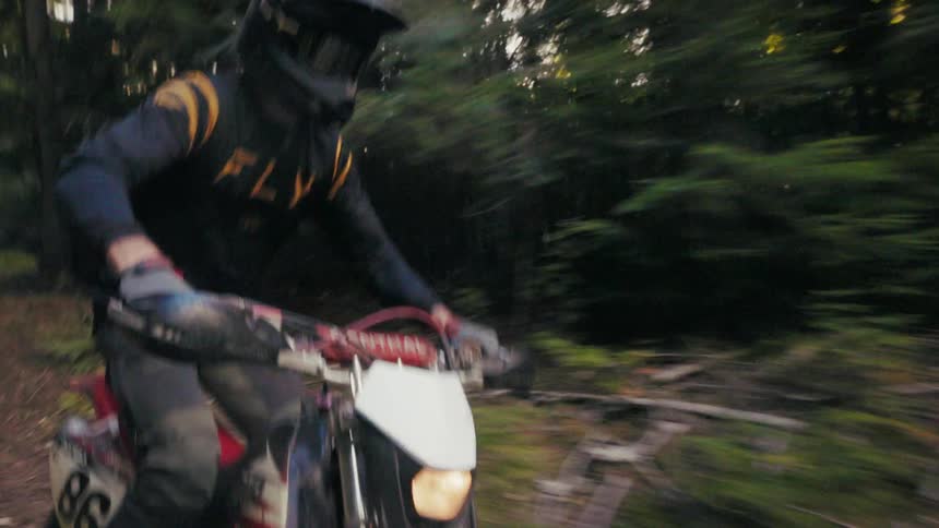 A man wearing a black helmet and a dark long sleeve jersey with gold stripes rides a motorcycle quickly on a dirt path through a dense forest with sunlight filtering through the trees
