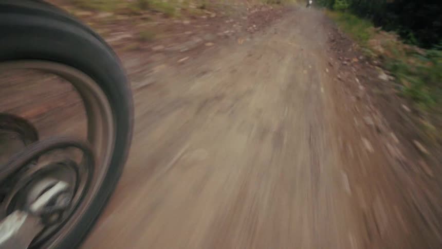 A low angle shot shows the rear wheel of a motorcycle spinning rapidly while traveling along a dirt path in a forest