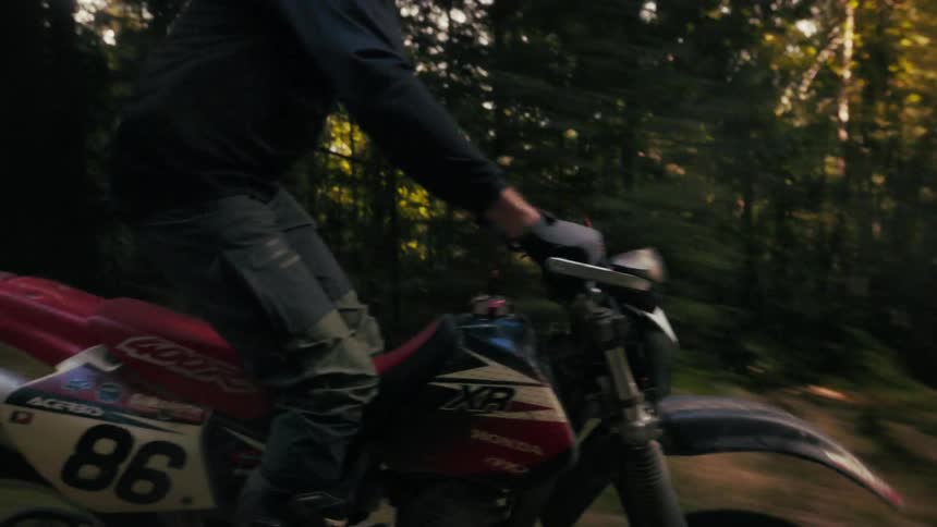 A rider in full protective gear maneuvers a dirt bike along a rugged treelined trailthe low sun filters through the forest canopy casting dynamic shadows as the motorcycle speeds across the uneven ground