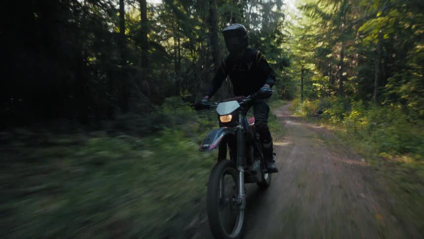 A motorcyclist wearing full black protective gear and a helmet rides a dirt bike down a narrow sundappled forest paththe camera follows from a low angle as the rider moves from bright sunlight into the deep shadows of the towering evergreen trees