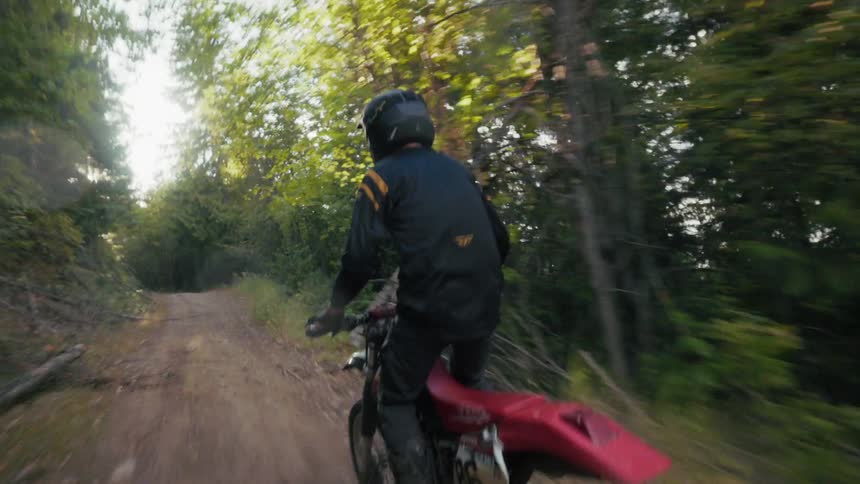 A motorcyclist rides along a dirt path through a sun dappled forest
